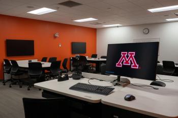 An instructor podium with a keyboard and monitor that is displaying the 'M' logo for the University of Minnesota is in the foreground. In the background there are computer monitors mounted on an orange wall with a table and chairs.