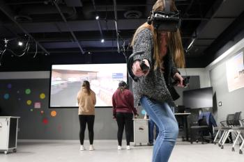 A student in the Virtual Reality Studio reaches one arm out in the direction of the camera. They are wearing a black headset and holding a controller in either hand. In the background, two more students look at a simulated grocery store on a projector screen. One student is wearing a headset.