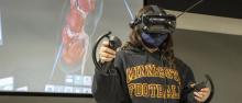 A student wearing a virtual reality helmet and hand controllers demonstrates a medical procedure on the screen behind them.