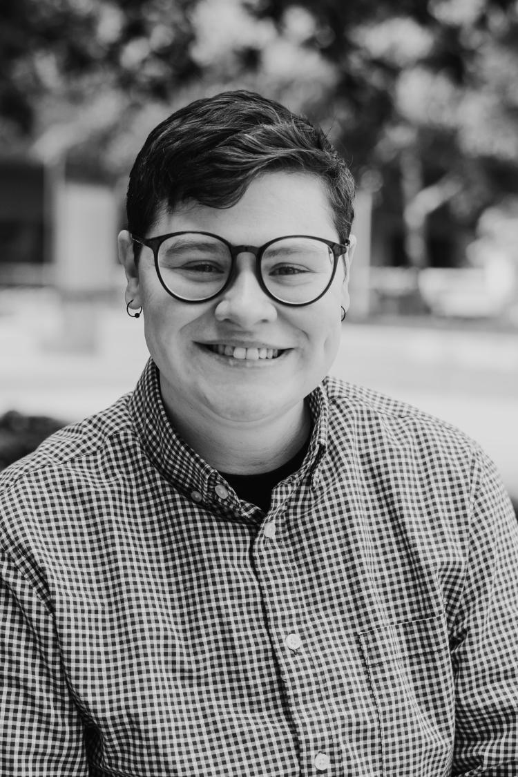White nonbinary person with short brown hair and glasses, smiling, and wearing a patterned button-up shirt