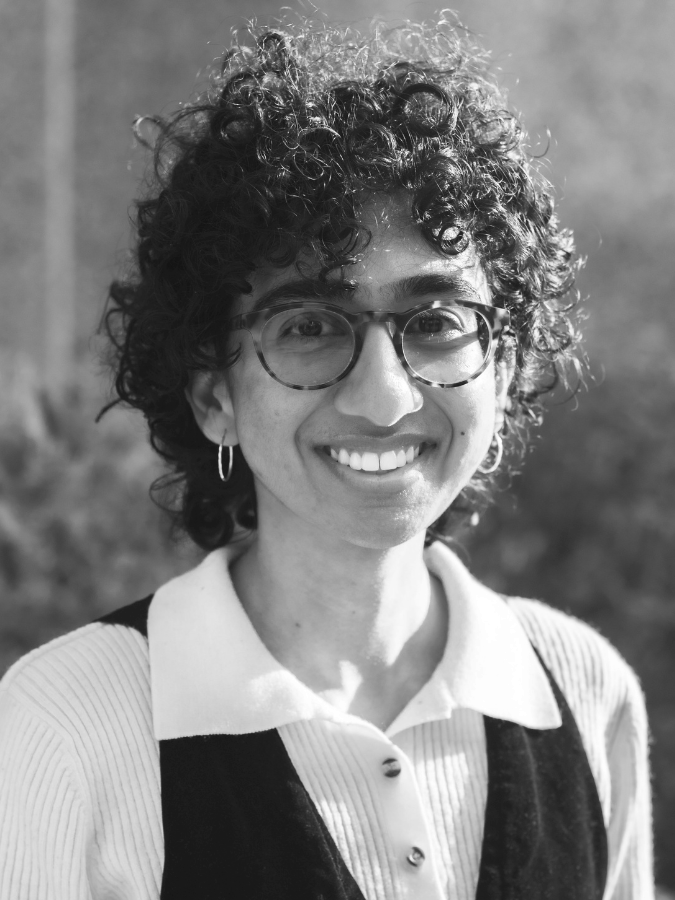 Headshot of Sana, a non-binary Indian person with curly hair and glasses. They are outside and smiling